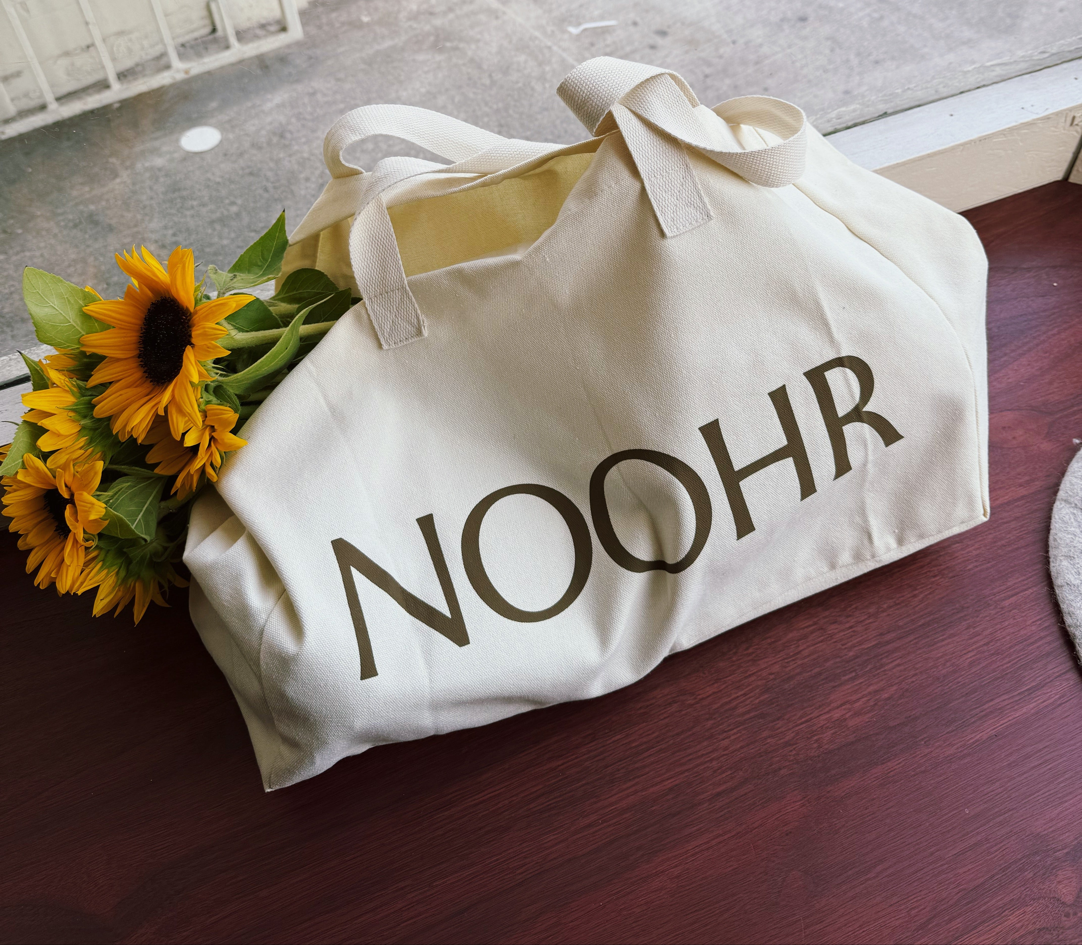 A cream canvas tote bag with the brand name 'NOOHR' printed in dark brown across the front, resting on a dark wooden table next to a bouquet of yellow sunflowers. The bag is positioned near a window looking out onto a city sidewalk.